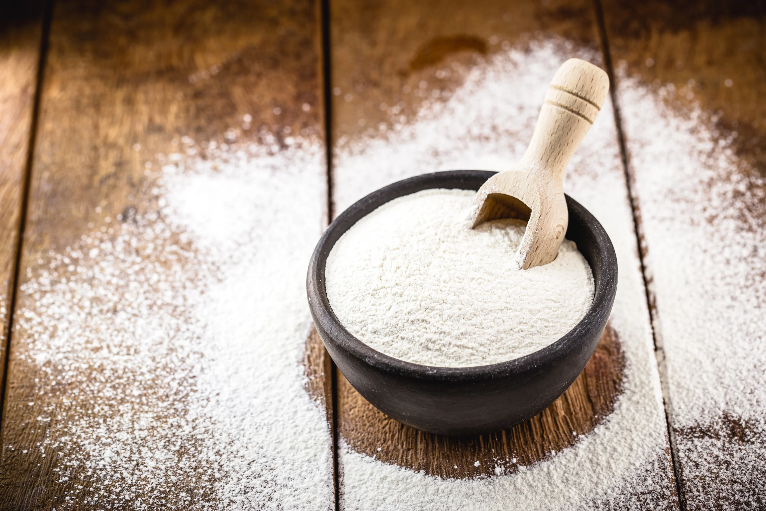 A brown bowl filled with white tapioca fiber flour sits on a wooden surface, with a small wooden scoop and loose flour scattered around.